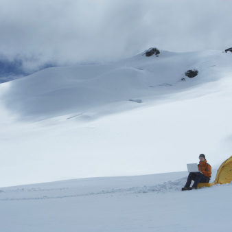 Hiker Using Laptop By Tent On Snowy Mountain Slope